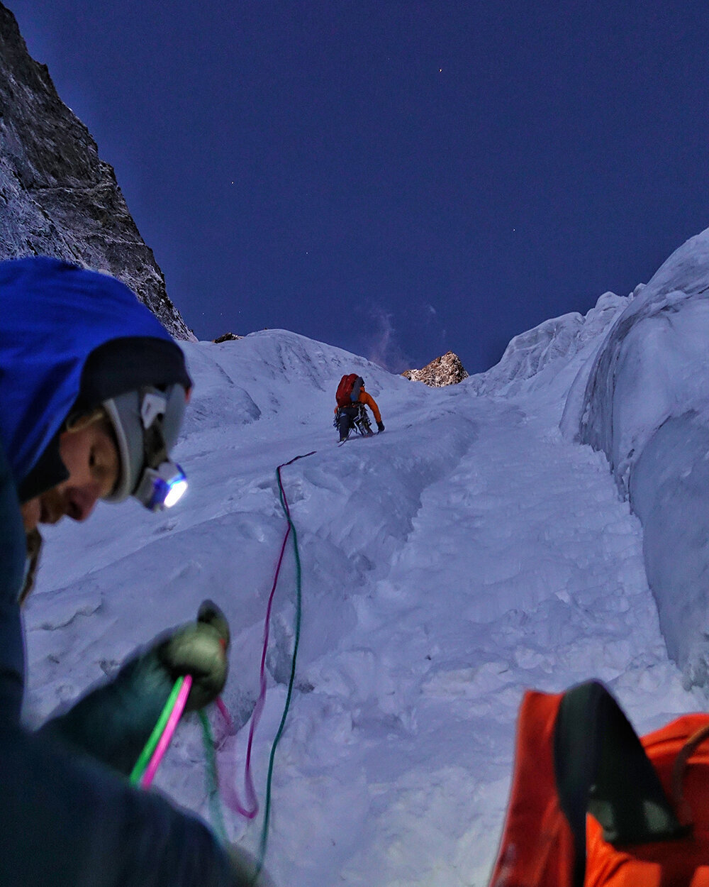 A belayer pays out rope as a climber above them climbs an icy couloir towards a starry sky.