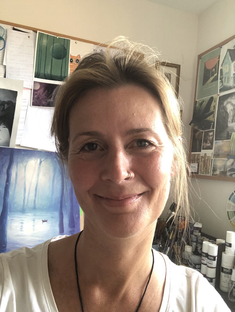 A woman with light brown hair in art studio with background art images on wall